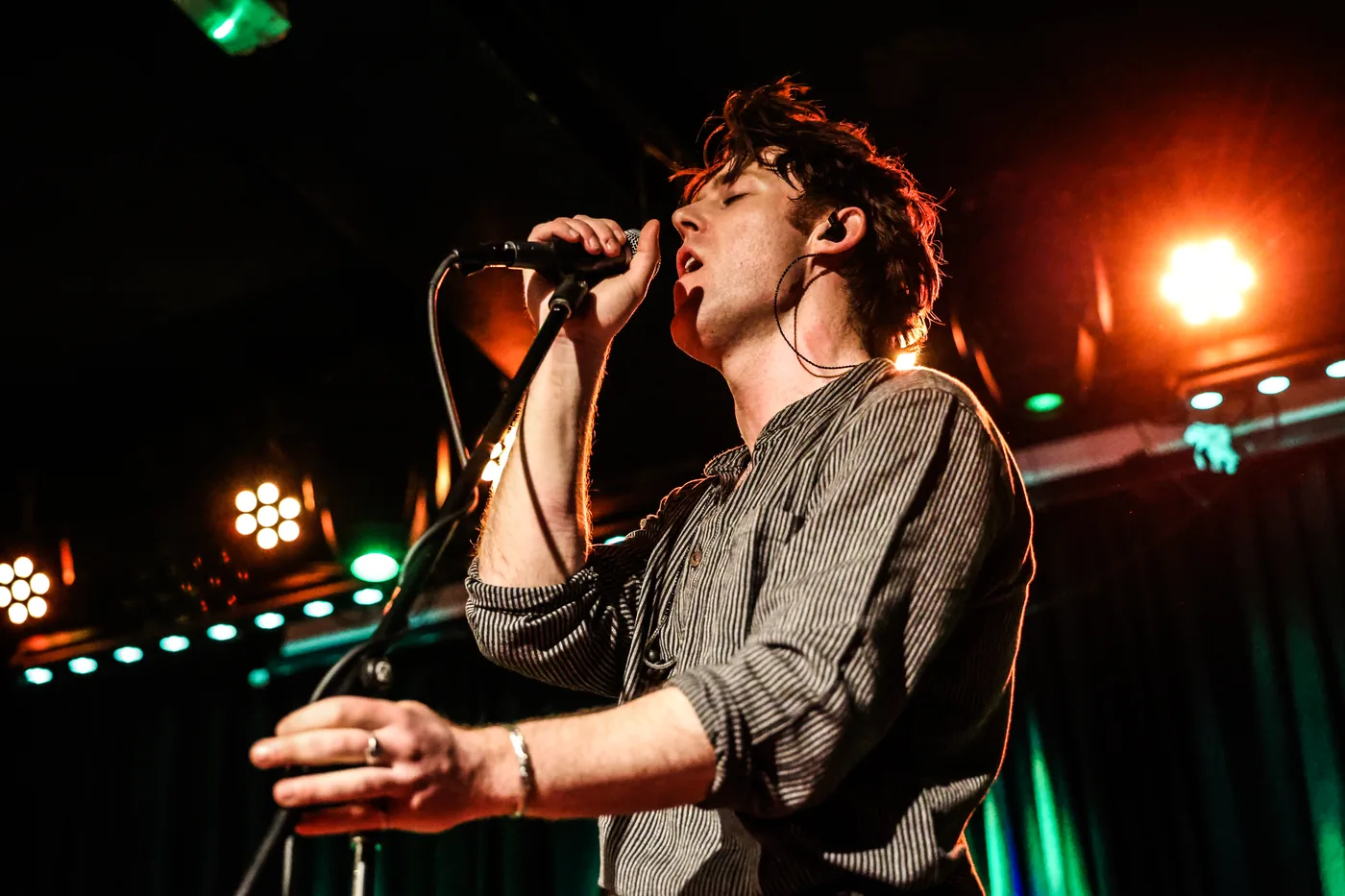 A singer leaning into the microphone with both hands, orange stage lights behind him