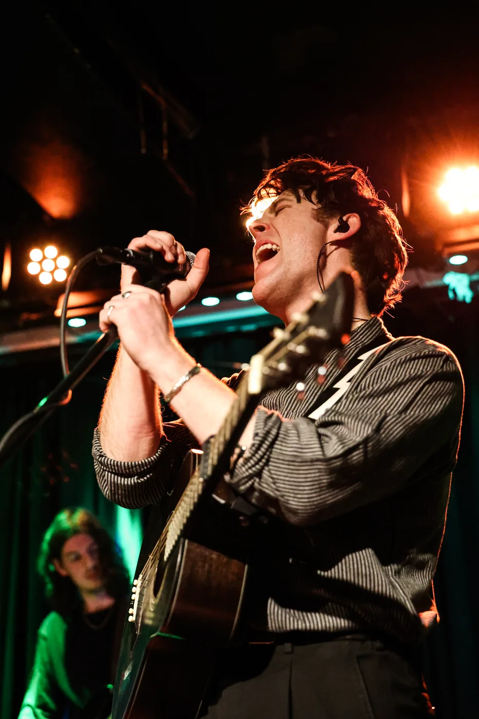 A singer gripping a microphone and singing, an acoustic guitar slung across his chest, orange stage lights behind