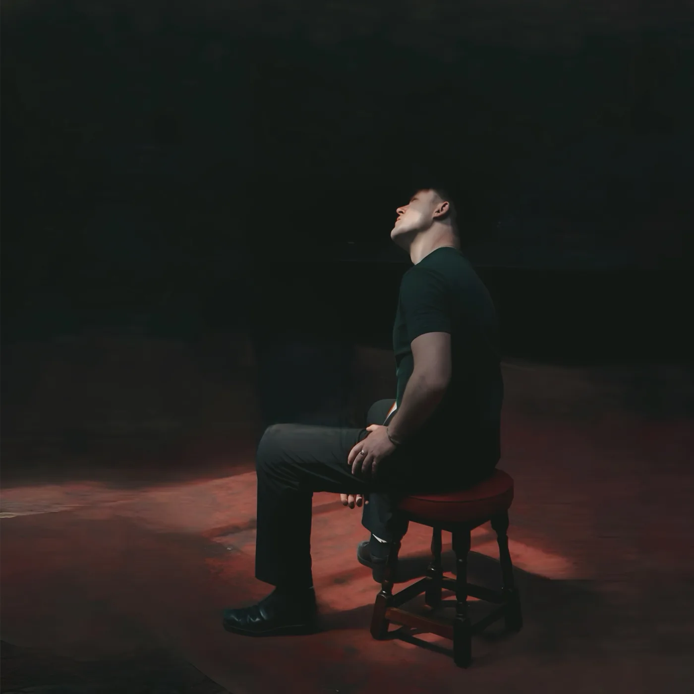 A man sitting alone on a wooden stool in the middle of a dark stage under a single light
