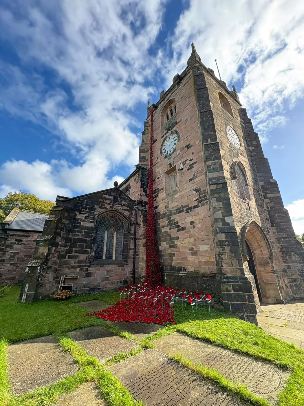 A long cascade of red poppies running from high on the stone clock tower of St Mary's Church, Radcliffe, down to a bed of poppies and small crosses on the grass